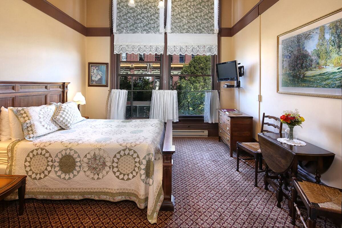 A hotel room with a bed, a desk, a chair, a TV, and a painting on the wall.