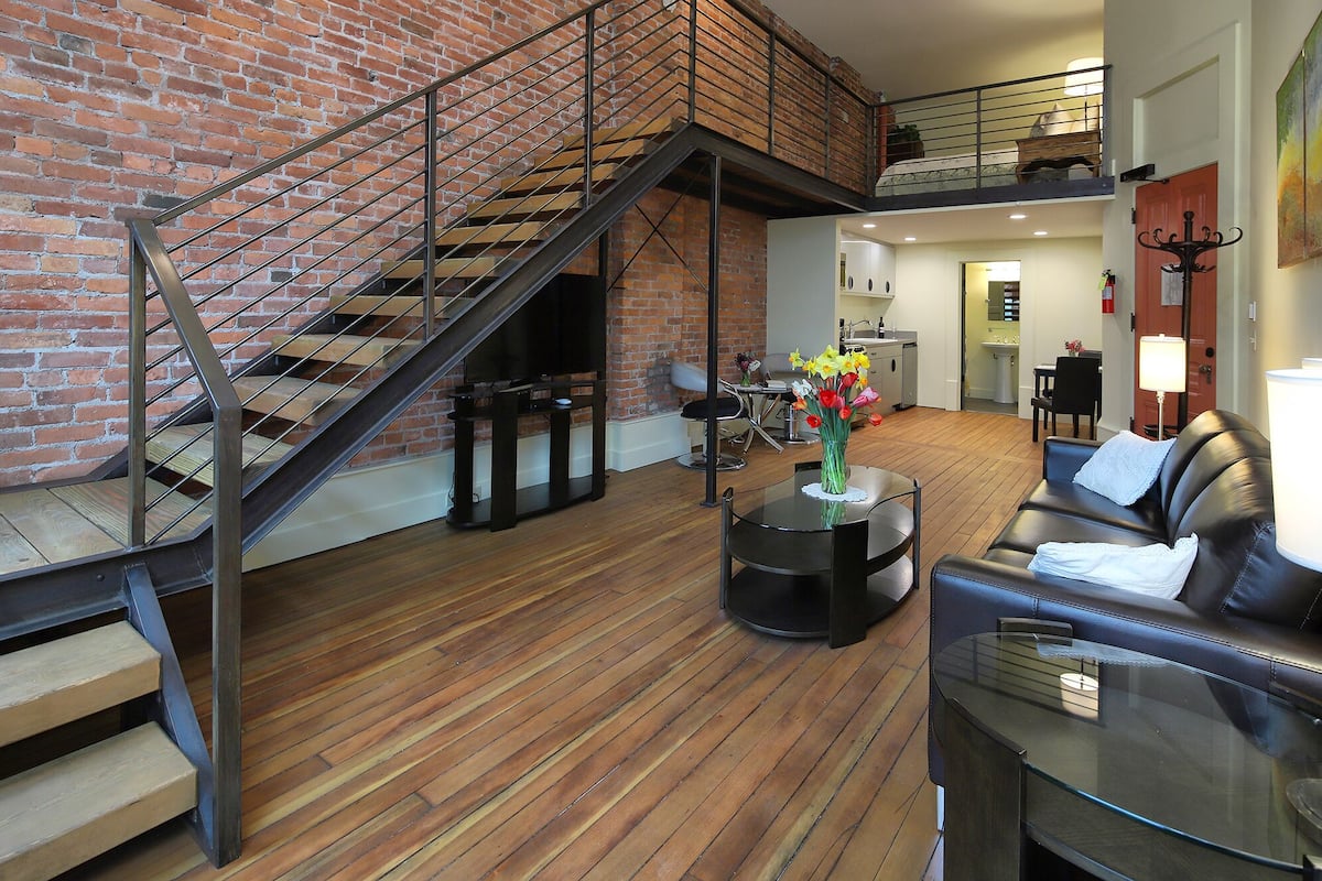 A spacious living area with a wooden floor, a black leather sofa, a glass coffee table, and a staircase with a metal railing.