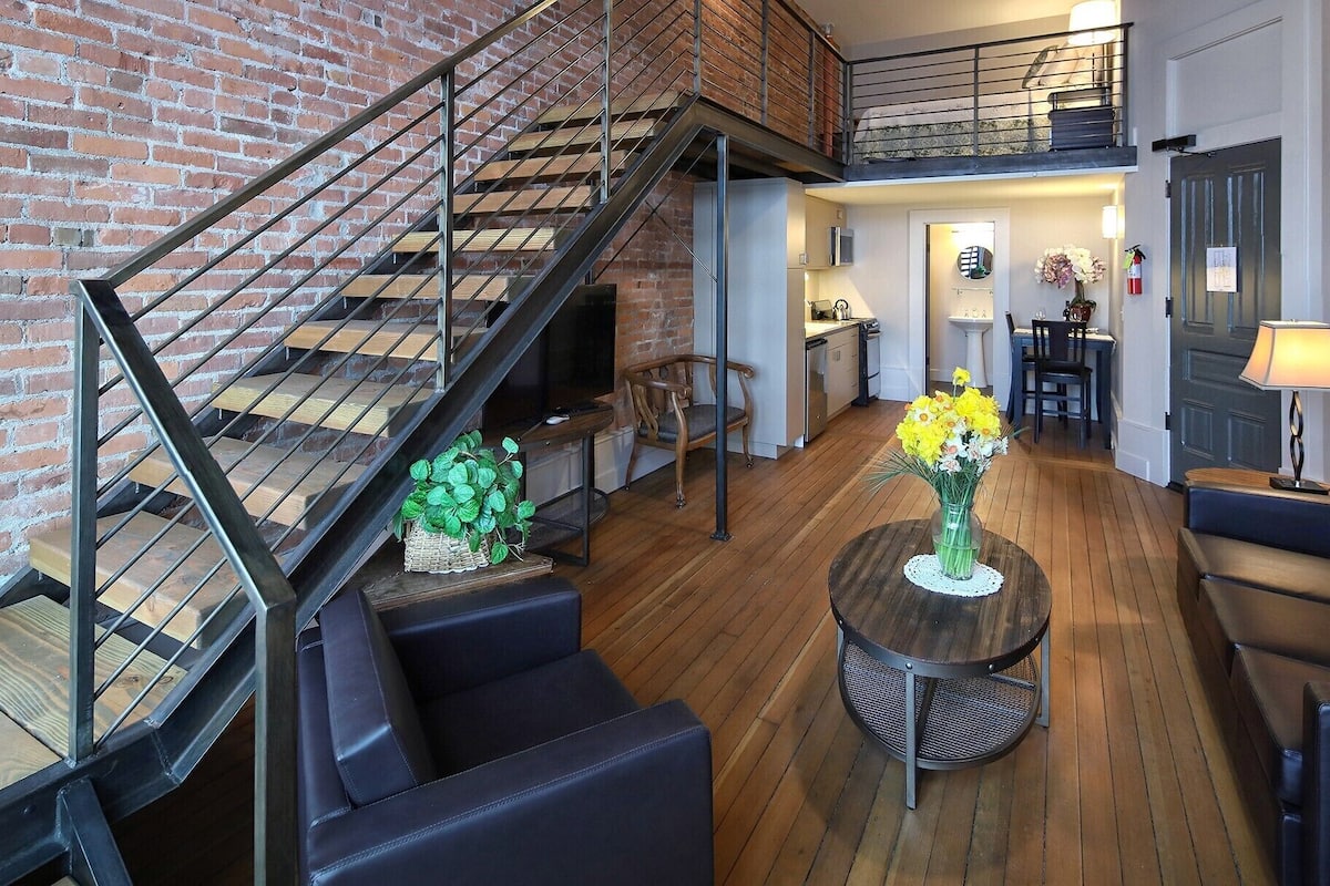 A loft-style living area with a wooden floor, a staircase with metal railings, a dining area with a table and chairs, and a seating area with sofas and a coffee table.