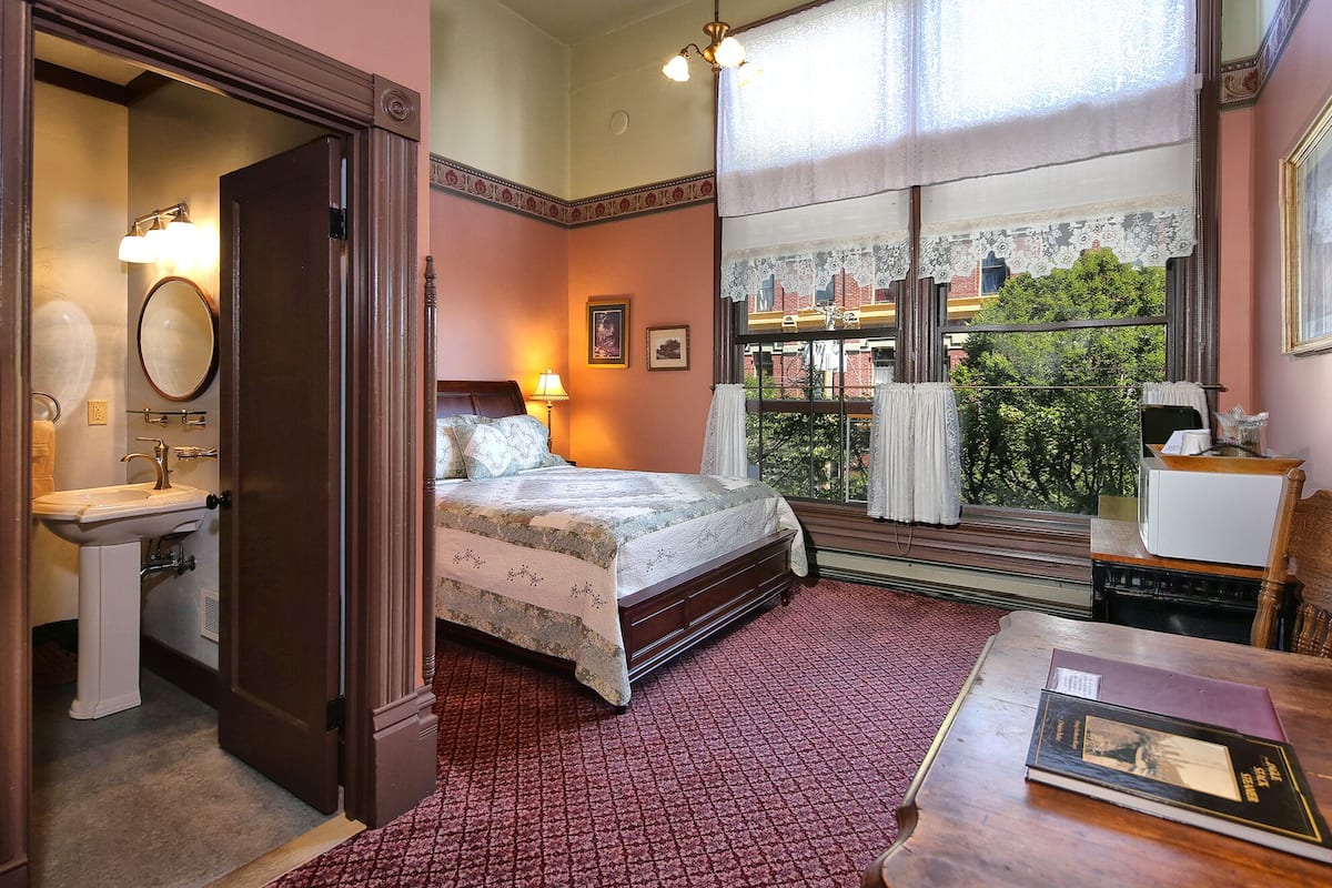 A bedroom with a bed, a bedside table with a lamp, a window with a view of buildings, and a door leading to a bathroom.