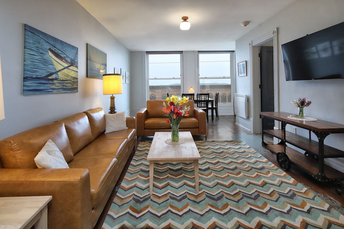 A living room with a brown leather sofa, a coffee table with a vase of flowers, a patterned rug, and a painting of a boat on the wall.