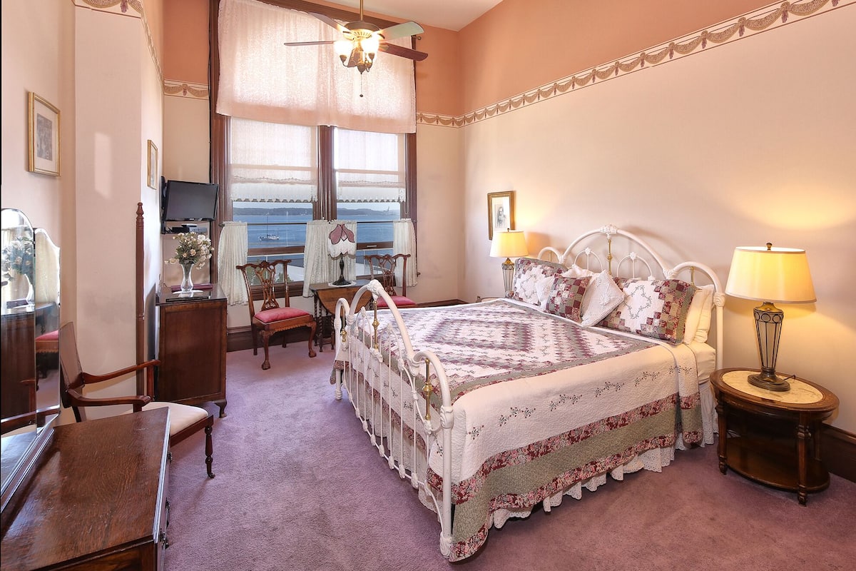 A bedroom with a large bed, two bedside tables with lamps, a chair, a desk, and a window with a view of the sea.