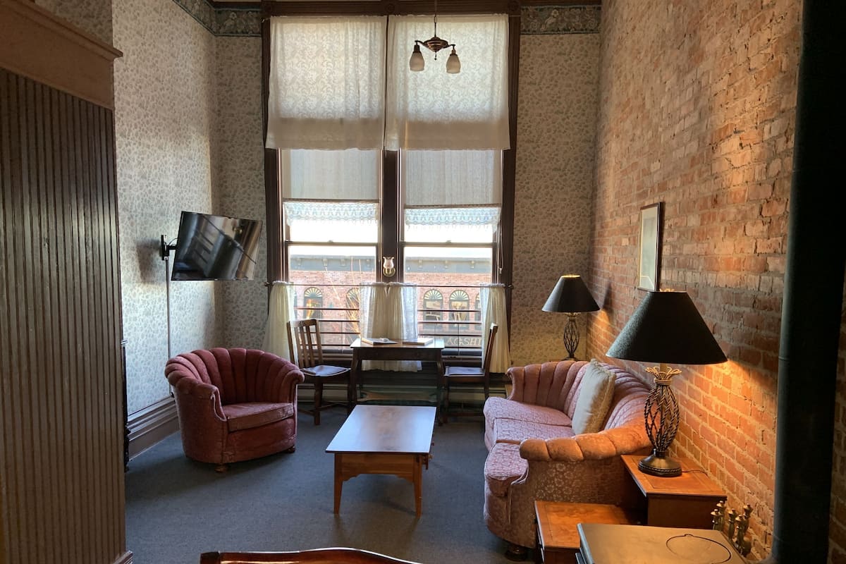 A cozy living room with a brick wall, a wooden table, two armchairs, a sofa, and a TV mounted on the wall.