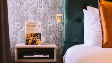 Un libro titulado 'A Century of Swansea' sobre una mesita de noche junto a una cama con cabecera verde y almohadas blancas.