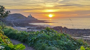 Un paysage de coucher de soleil sur la côte, avec un phare, un terrain rocheux et de la végétation.