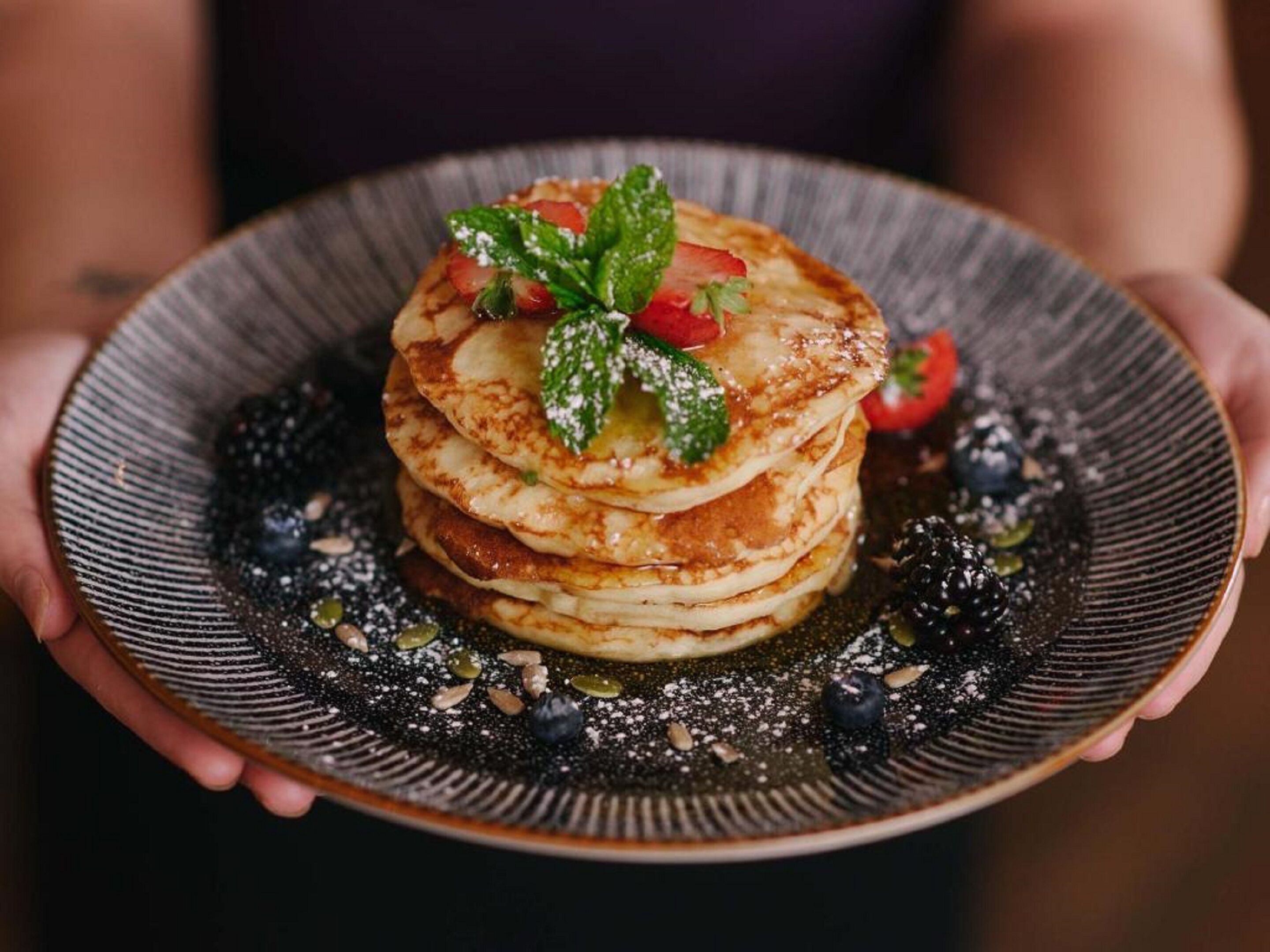 A stack of pancakes on a plate with a dark rim, garnished with mint leaves and a strawberry.