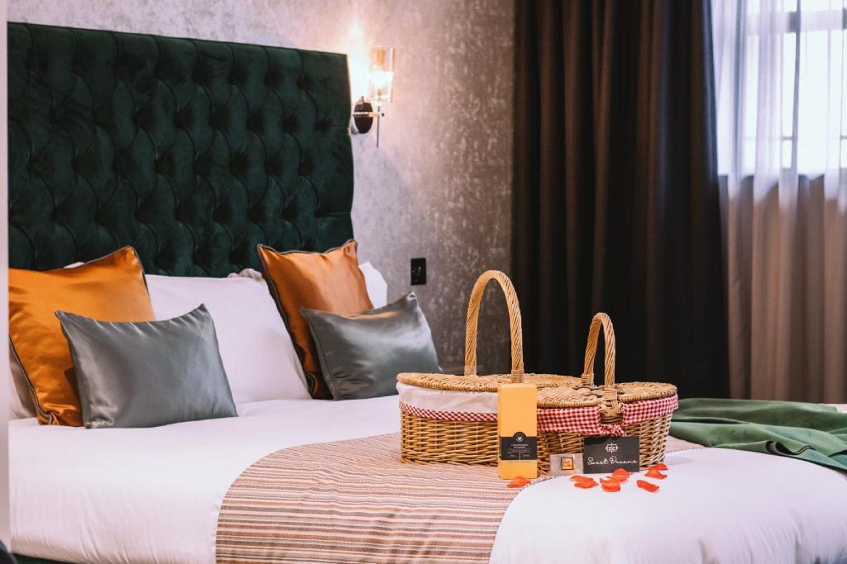 Een opgemaakt bed met een gestreept dekbedovertrek, een donkergroen gestoffeerd hoofdbord en een rieten mand met een label.