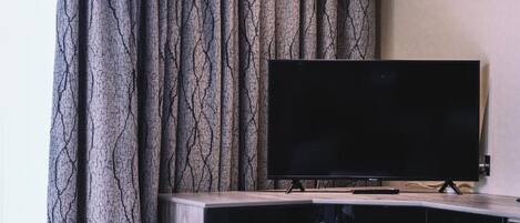 A flat-screen TV on a wooden cabinet with a remote control, set against a curtain with a tree-like pattern.