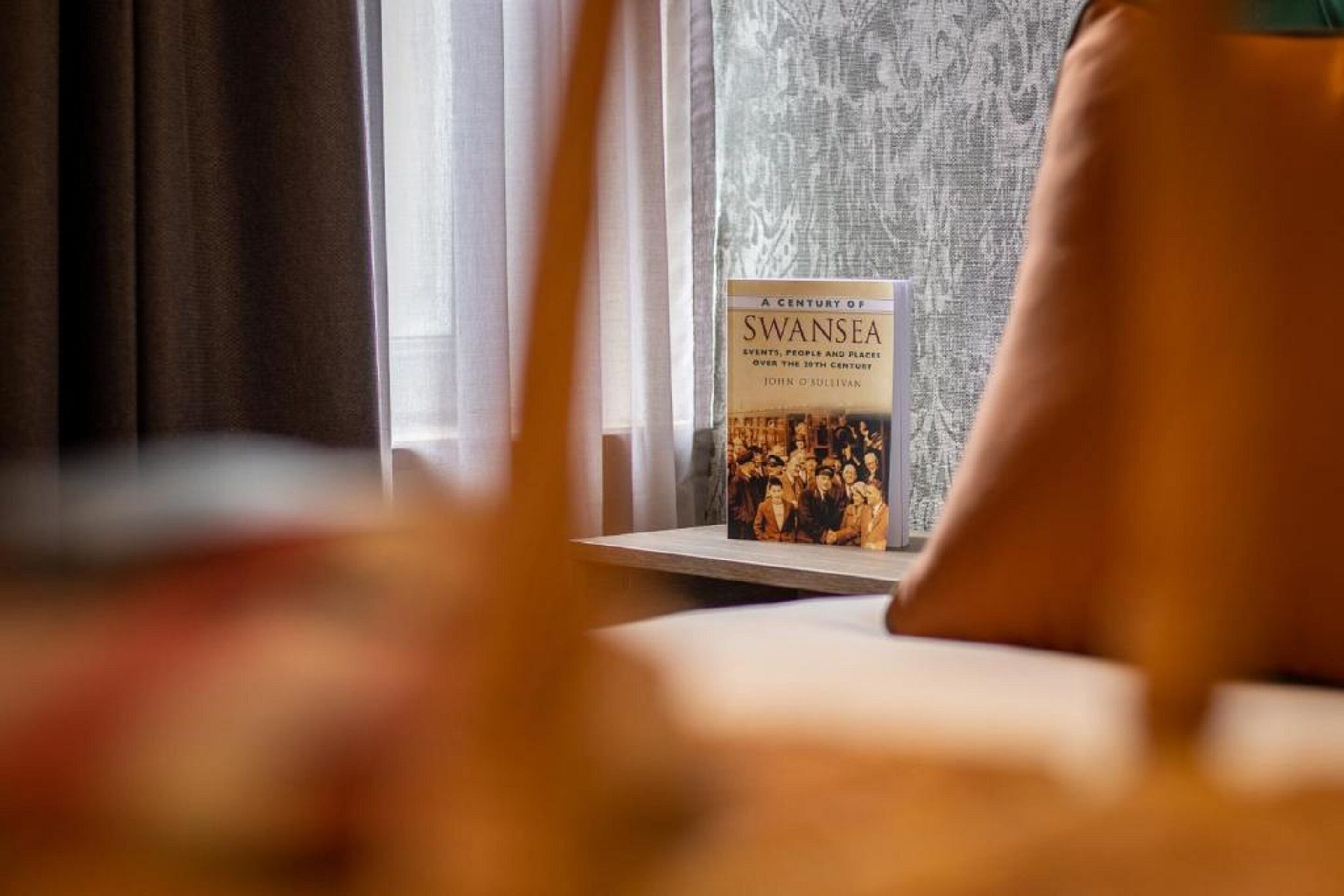 Un libro titulado 'A Century of Swansea' en una estantería cerca de una ventana con cortinas.