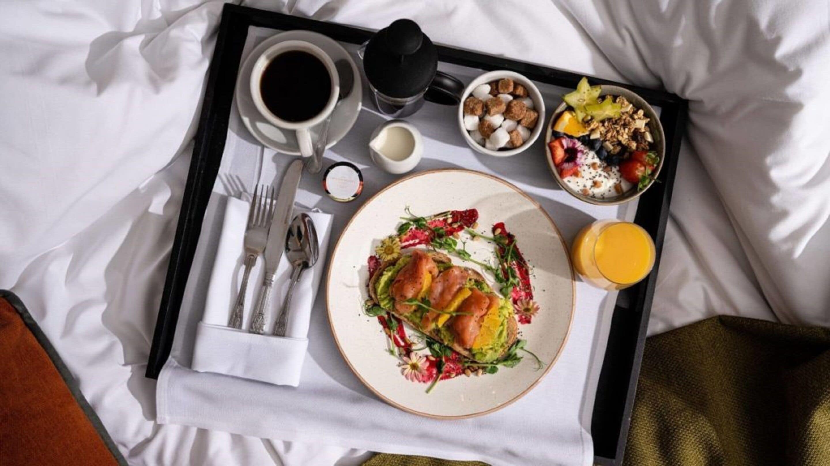 A breakfast tray with a plate of food, coffee, a juice glass, and a small bowl of cereal on a bed.