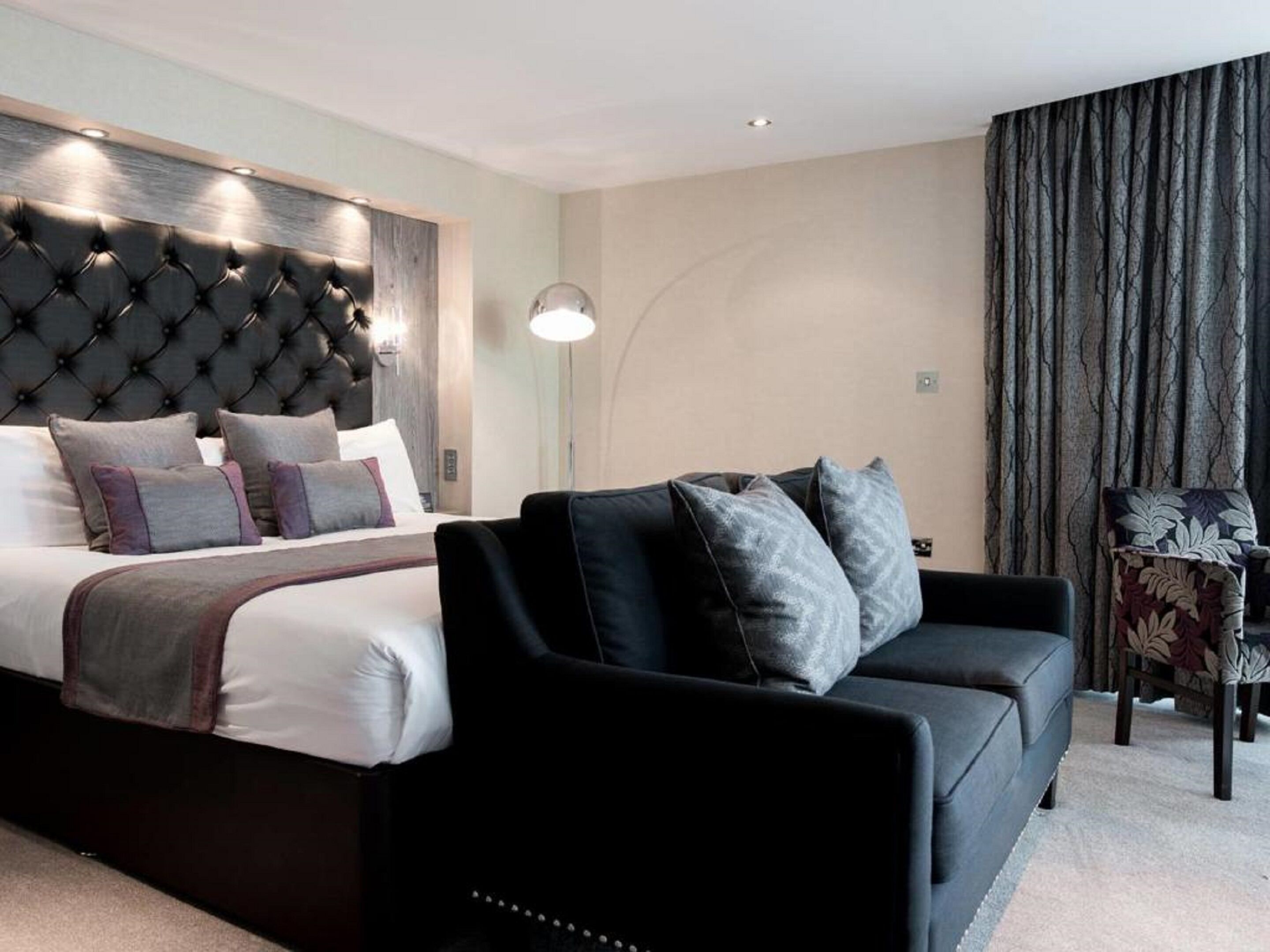 A modern hotel room with a large bed, a black sofa, and a small armchair.