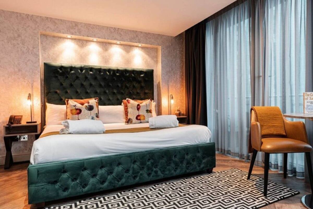 Une chambre d’hôtel avec un grand lit, une tête de lit recouverte d’un tissu vert, un fauteuil en cuir marron, une petite table avec un livre et un tapis à motifs.