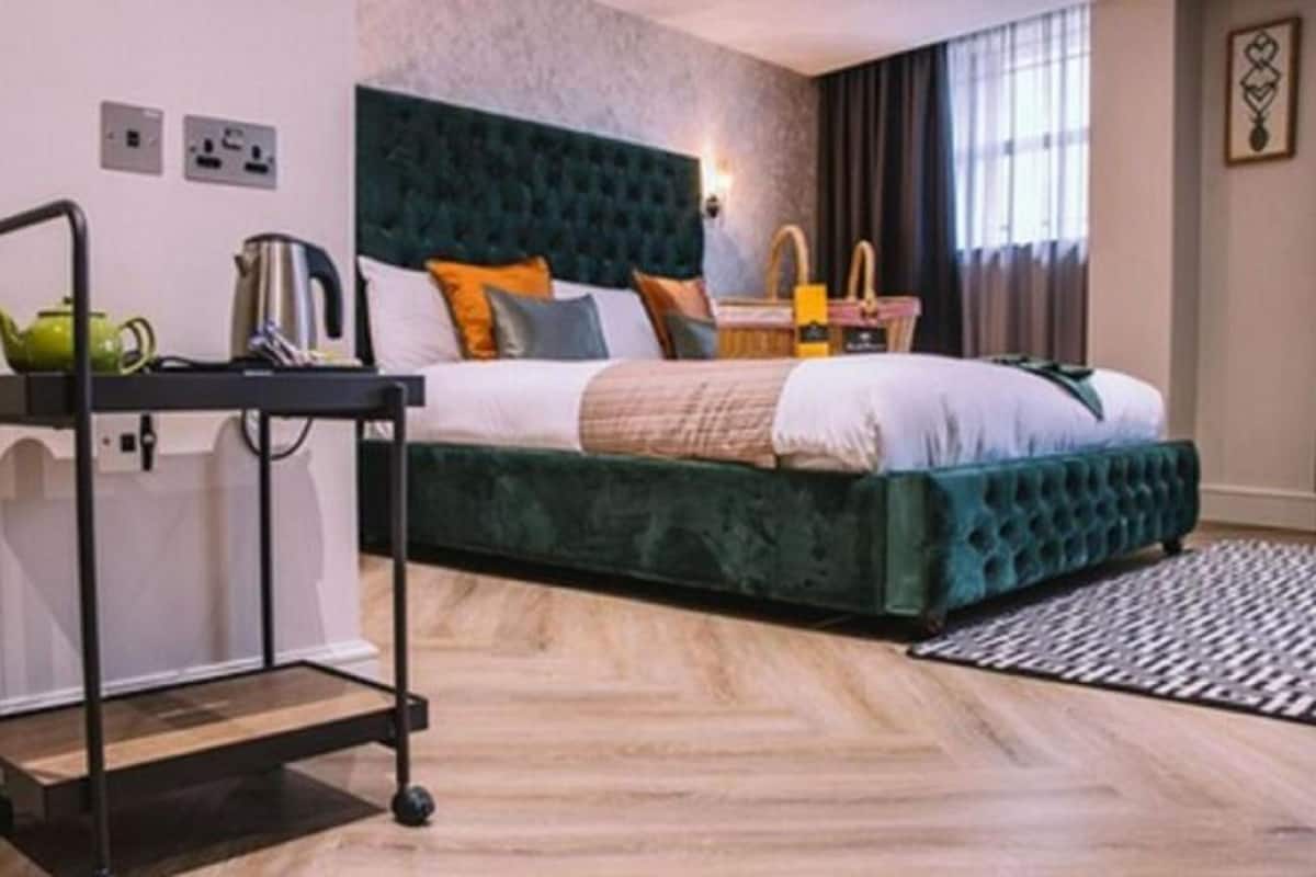 Une chambre d’hôtel avec un lit recouvert d’un tissu vert, une petite table sur laquelle se trouve une théière verte, un chariot en métal noir et une fenêtre avec des rideaux.
