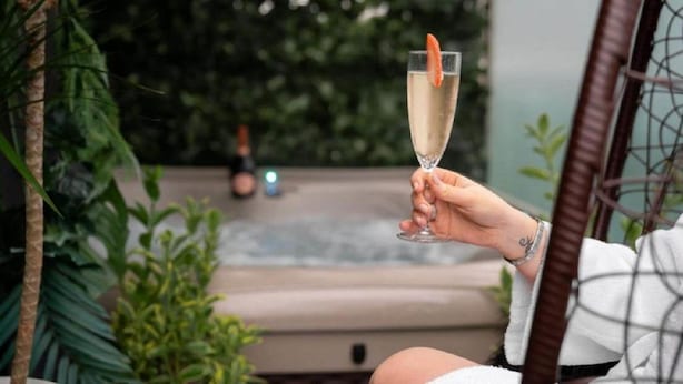 Une personne tenant un verre de champagne décoré d’une fraise, assise dans un fauteuil en osier, avec en arrière-plan un jacuzzi et de la verdure.