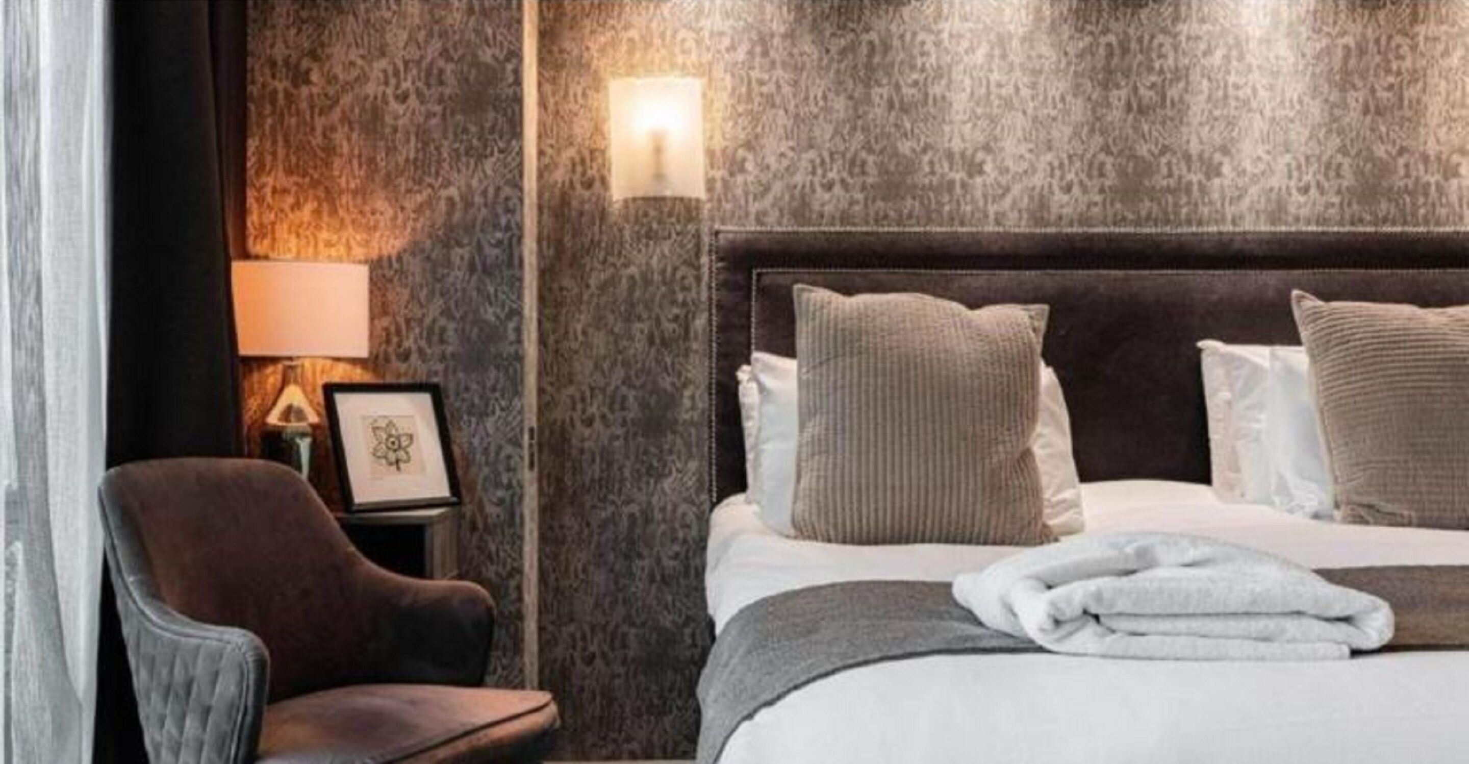 Ein Hotelzimmer mit einem großen Bett, einem dunklen Kopfteil, einem braunen Sessel, einem gerahmten Bild auf dem Nachttisch und einer Wandlampe.