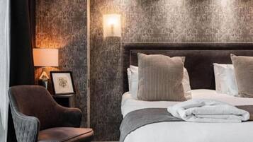 Quarto de hotel com uma cama grande, cabeceira escura, poltrona marrom, um quadro na mesa de cabeceira e uma luminária fixada na parede.