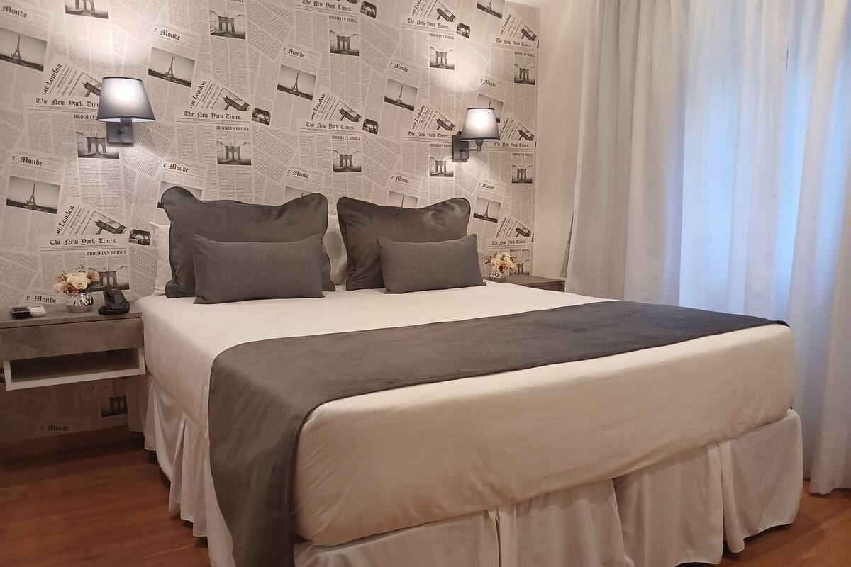 A neatly arranged bedroom with a large bed, two bedside lamps, a nightstand with a phone and a vase, and a wall covered with newspaper clippings.