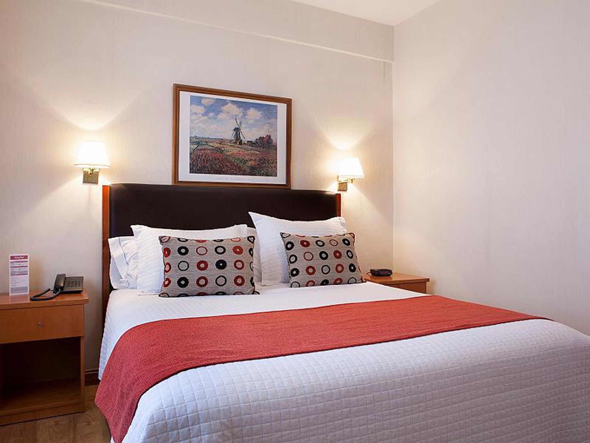 A hotel room with a bed, two bedside lamps, a framed picture on the wall, and a nightstand with a phone.