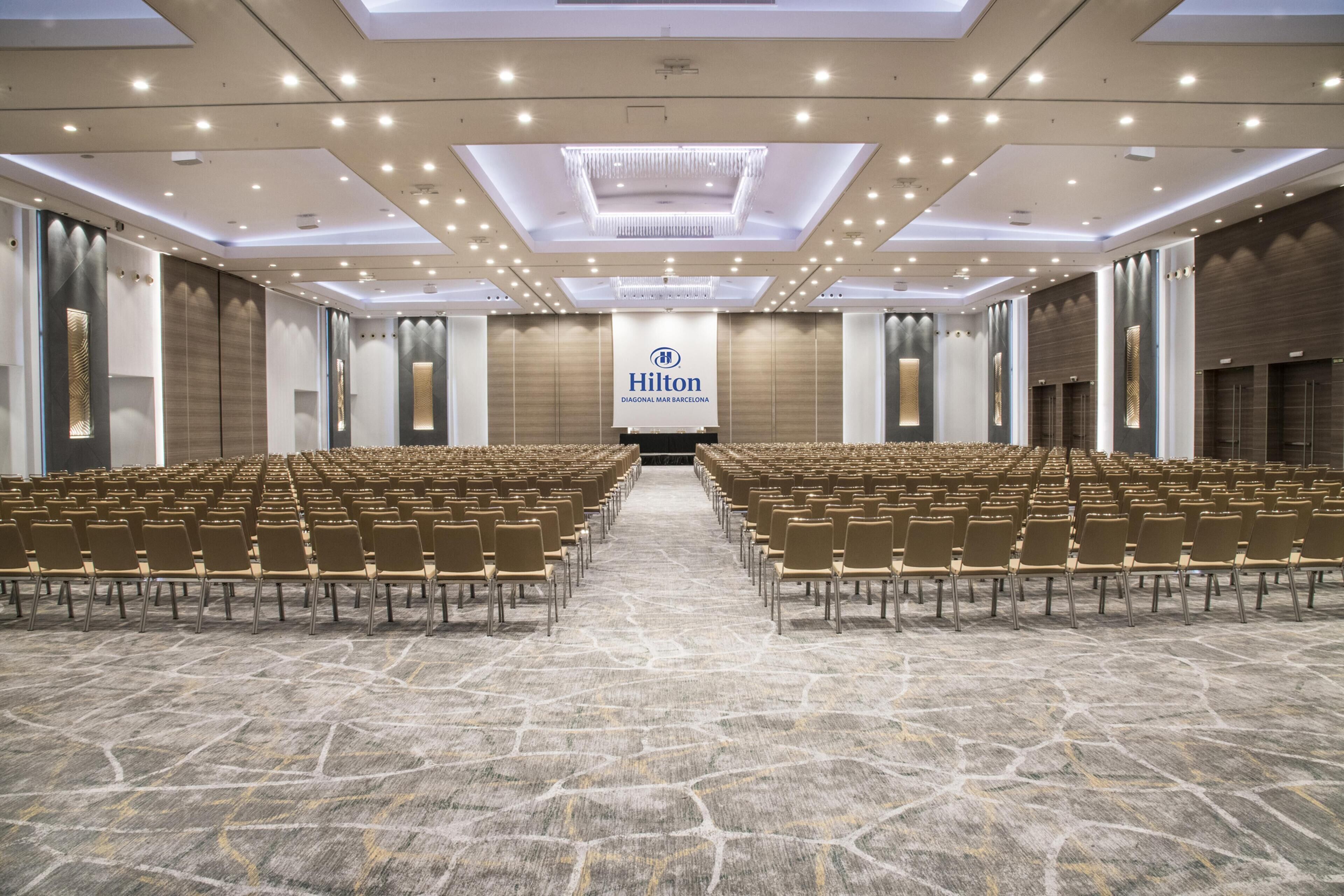 Una amplia sala de conferencias con filas de sillas, un gran cartel de Hilton y un moderno diseño de techo.