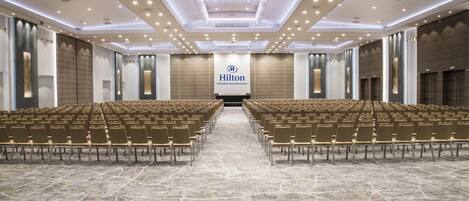 Una amplia sala de conferencias con filas de sillas, un gran cartel de Hilton y un moderno diseño de techo.