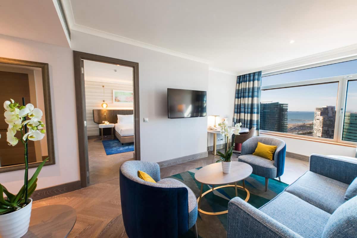A modern hotel room with a large window, a flat-screen TV, and a seating area with a coffee table and chairs.