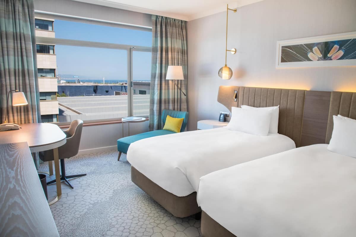 A hotel room with two beds, a desk, a chair, a small table, and a view of the cityscape.