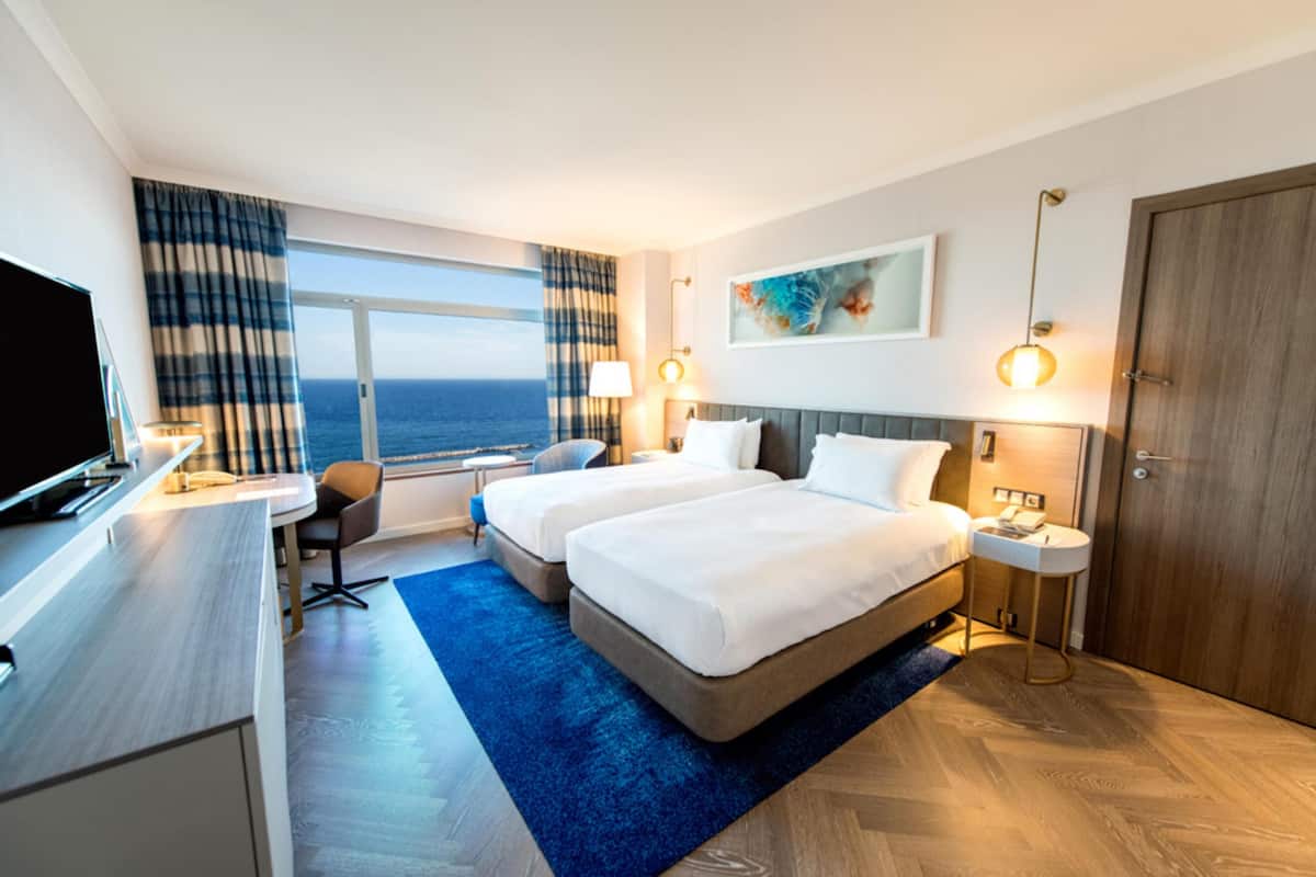 A hotel room with two beds, a desk, a TV, and a view of the ocean.