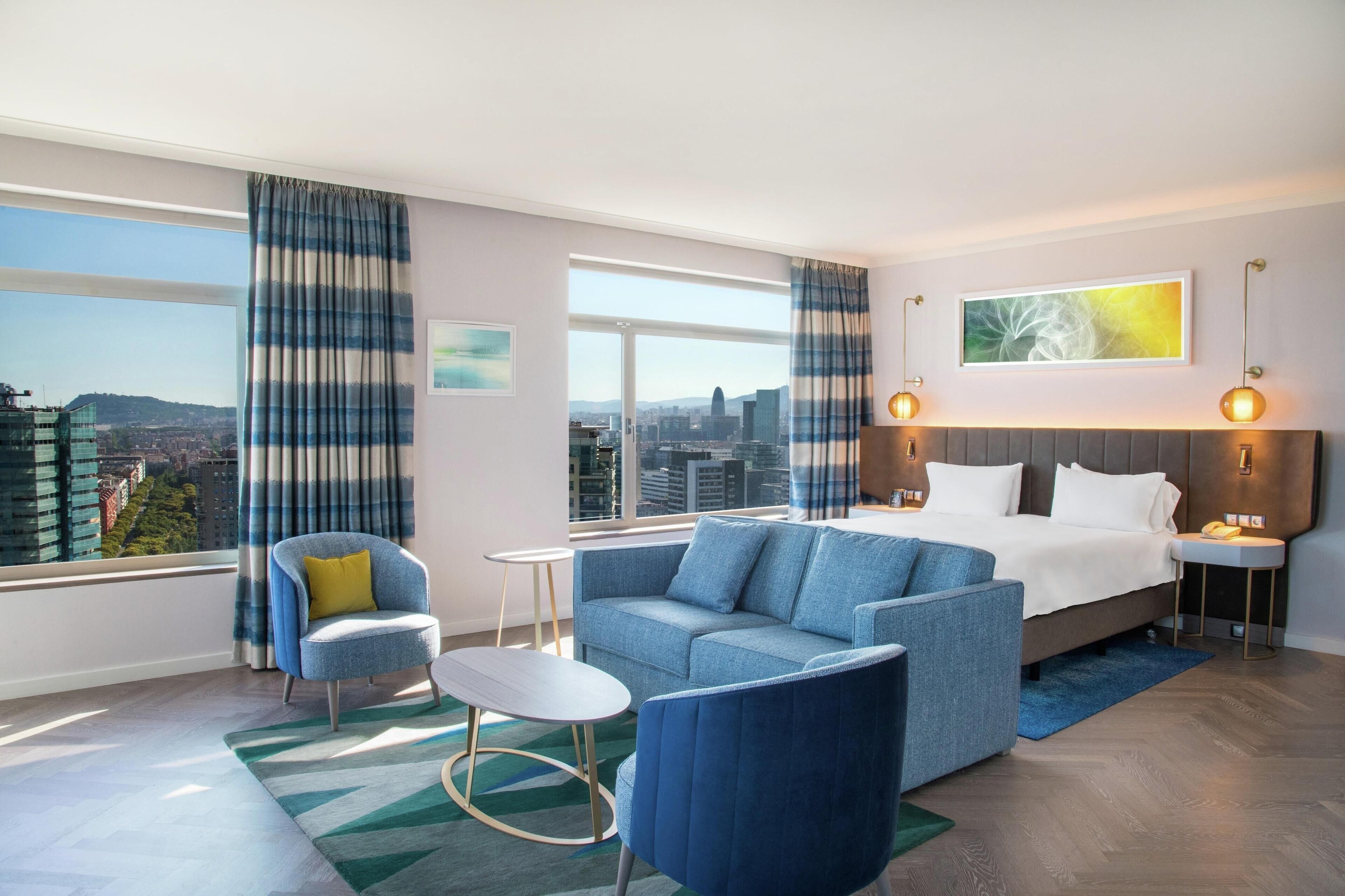 A modern hotel room with a large bed, a sofa, a chair, and a small table. There are large windows with curtains, a painting on the wall, and a city view.