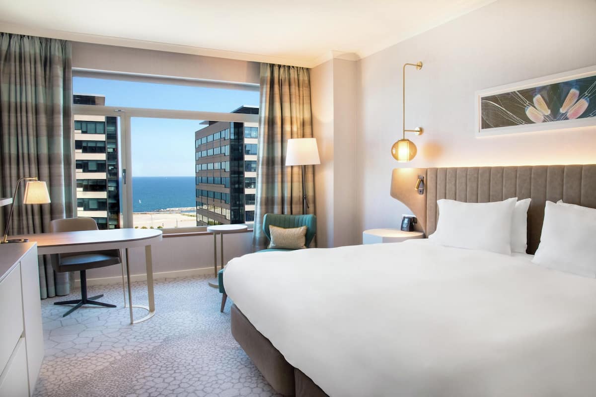 A hotel room with a large bed, a desk, a chair, a view of the ocean, and a cityscape.
