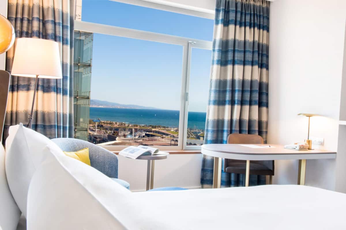 A hotel room with a bed, a small table, a chair, a lamp, and a view of the sea and cityscape through large windows.