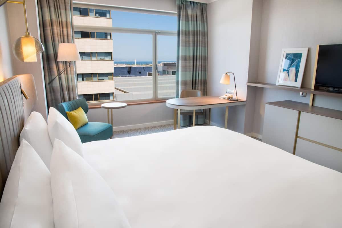 A hotel room with a large bed, a desk, a chair, a TV, and a window with a view of buildings and the sky.