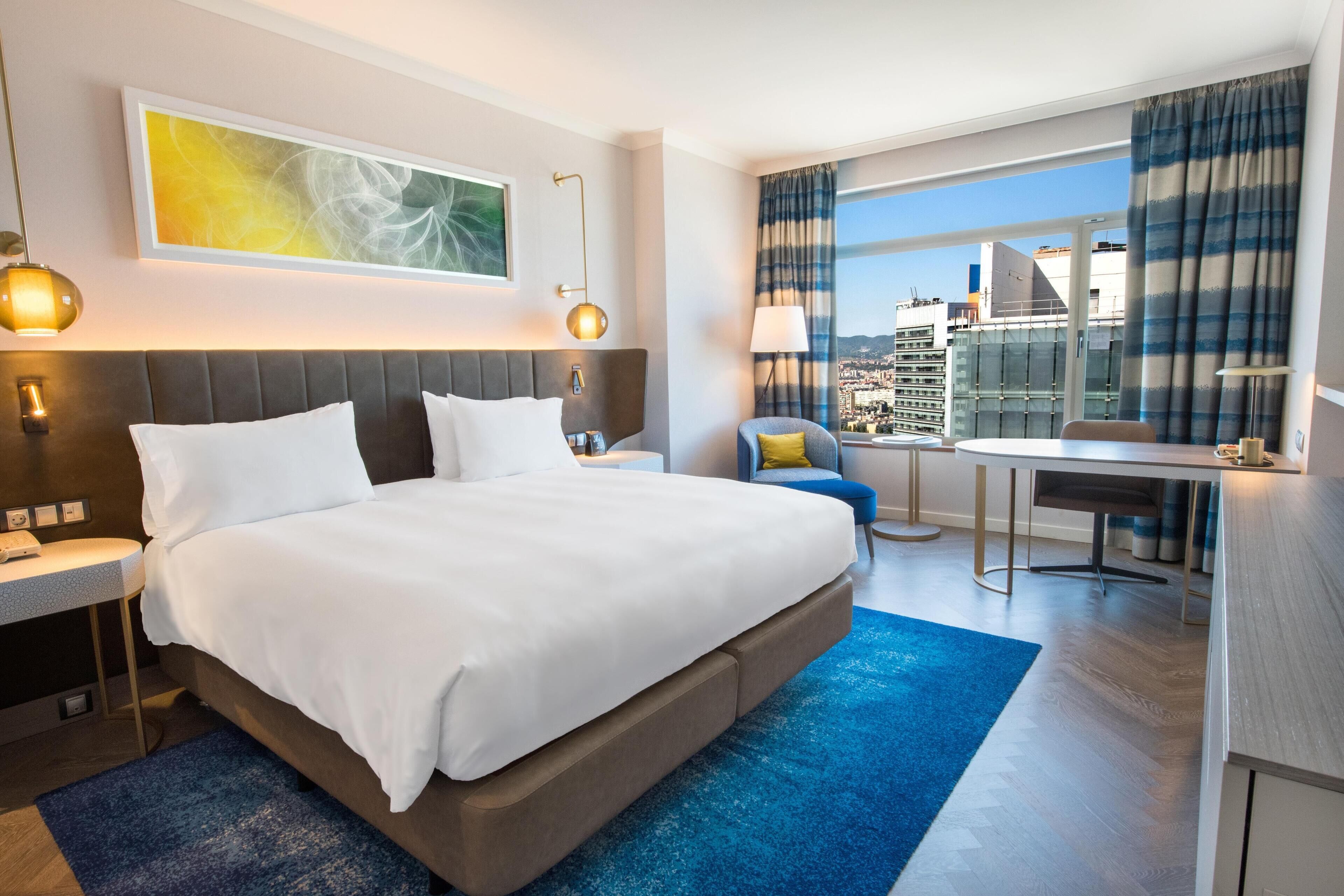 A modern hotel room with a large bed, a desk, and a view of the city.