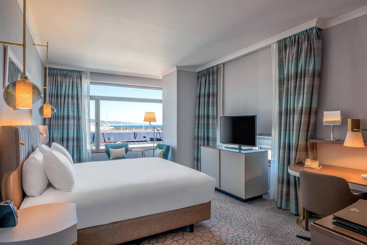 A hotel room with a large bed, a desk with a flat-screen TV, a dining area with a table and chairs, and a view of the cityscape through the window.