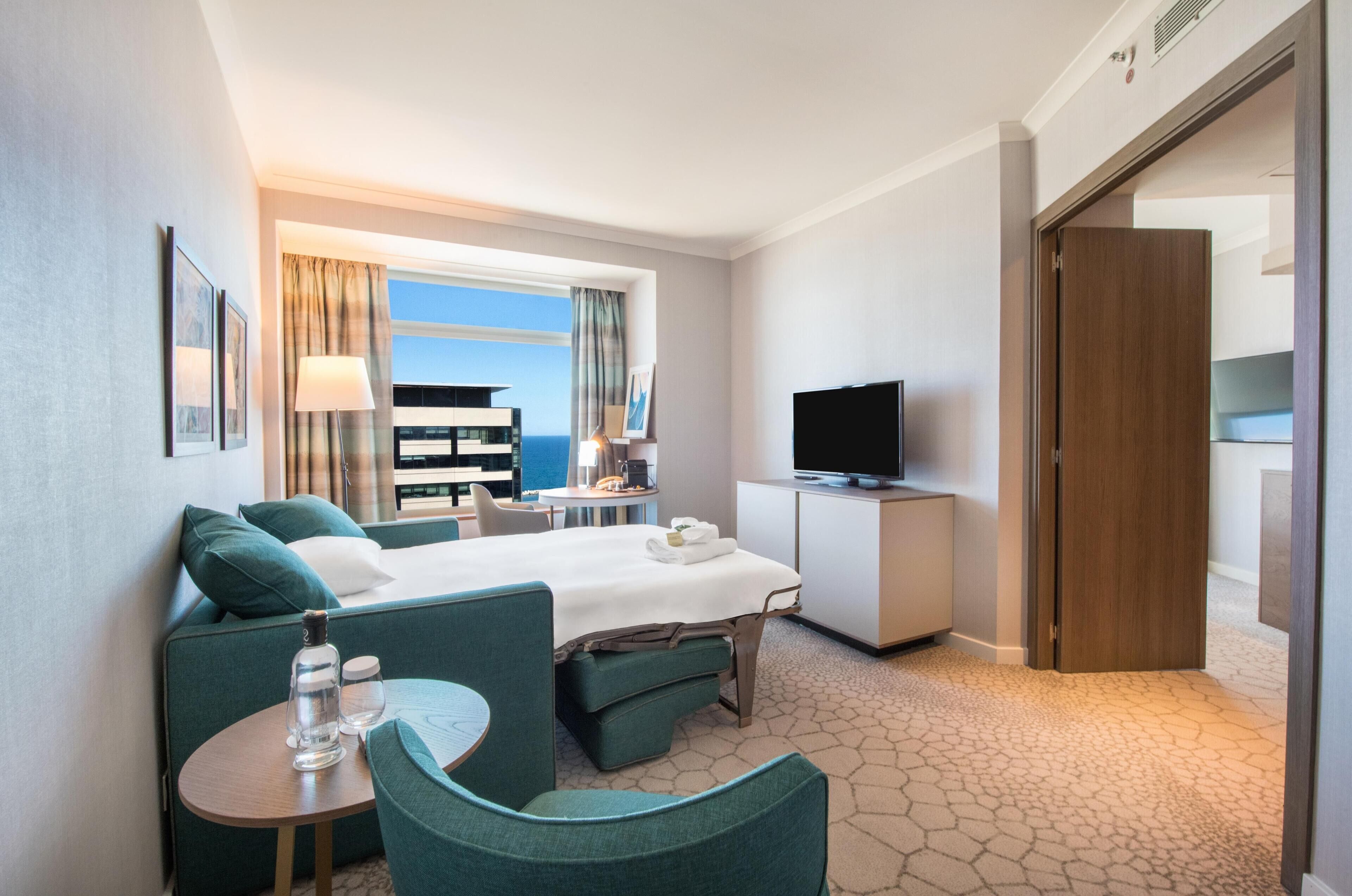 Habitación de hotel con cama, zona de estar, televisión y vista al océano.