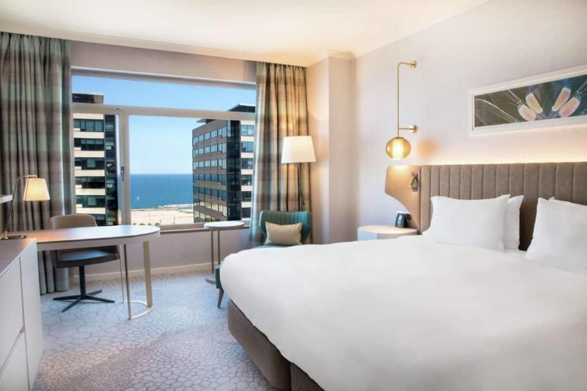 A hotel room with a bed, a desk, a chair, a lamp, and a view of the ocean.