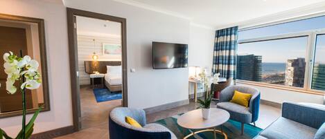 A modern hotel room with a large window, a flat-screen TV, and a seating area with blue and gray furniture.