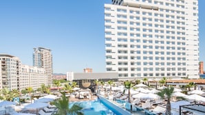 A rooftop pool area with lounge chairs, umbrellas, and a large building in the background.
