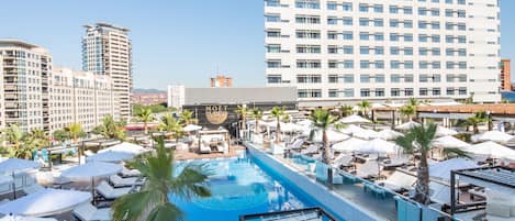A rooftop pool area with lounge chairs, umbrellas, and a large building in the background.