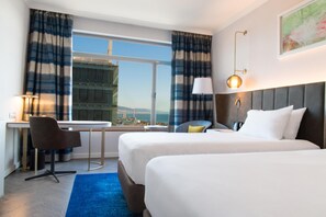 A hotel room with two beds, a desk, a chair, a lamp, and a view of a cityscape. - Hilton Diagonal Mar Barcelona (Barcelona)