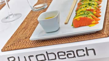 Un plato de sushi con un recipiente pequeño de salsa y palillos sobre un plato blanco, colocado sobre una mesa con un mantel individual que dice "Purobeach".