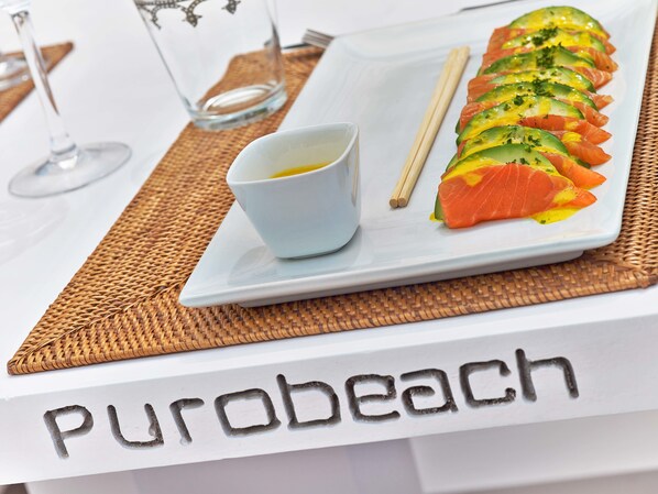 Un plato de sushi con un recipiente pequeño de salsa y palillos sobre un plato blanco, colocado sobre una mesa con un mantel individual que dice "Purobeach".