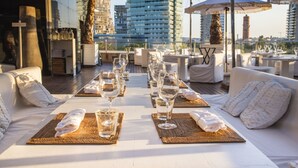 A rooftop dining area with white furniture, arranged tables with wine glasses, and a city skyline view.