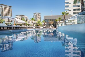 A swimming pool with clear blue water, surrounded by palm trees and modern buildings. - Hilton Diagonal Mar Barcelona (Barcelona)