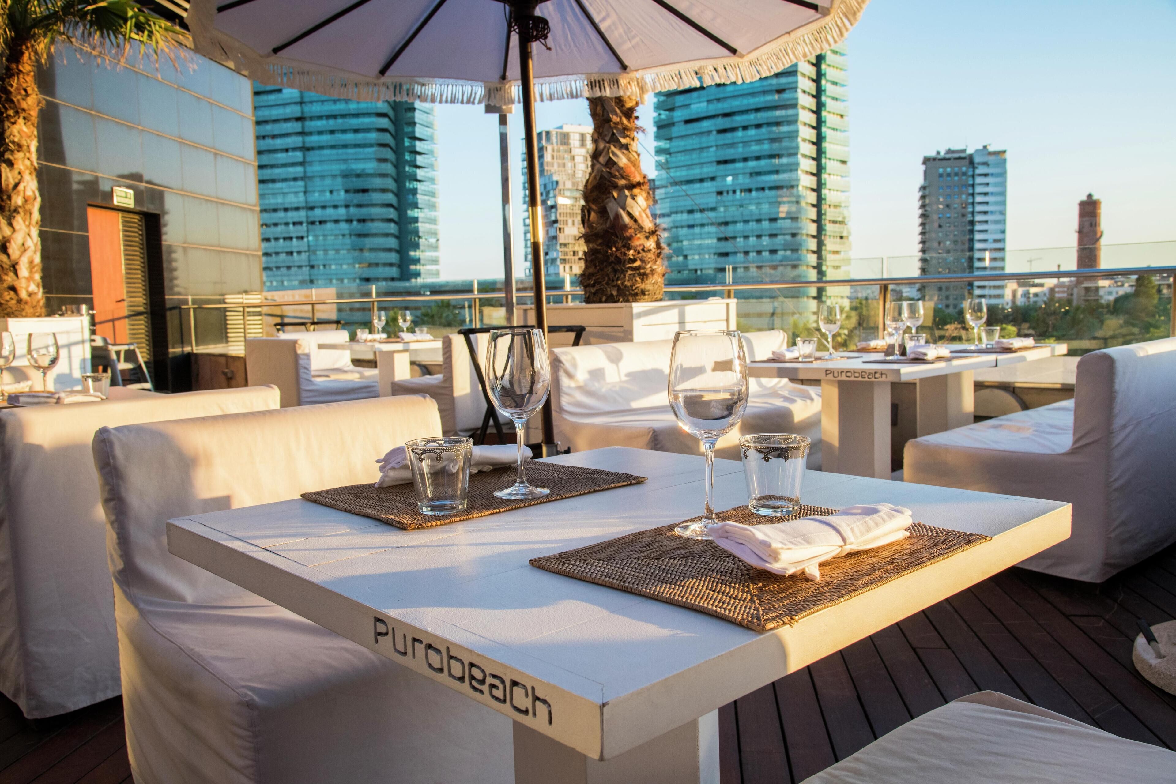 Un área de comedor en la azotea con mesas preparadas para dos, cada una con un vaso de agua y una copa de vino. Las mesas están cubiertas con manteles blancos y tienen individuales. Se ve el nombre 'Purobeach' en la mesa.