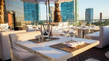 Un área de comedor en la azotea con mesas preparadas para dos, cada una con un vaso de agua y una copa de vino. Las mesas están cubiertas con manteles blancos y tienen individuales. Se ve el nombre 'Purobeach' en la mesa.