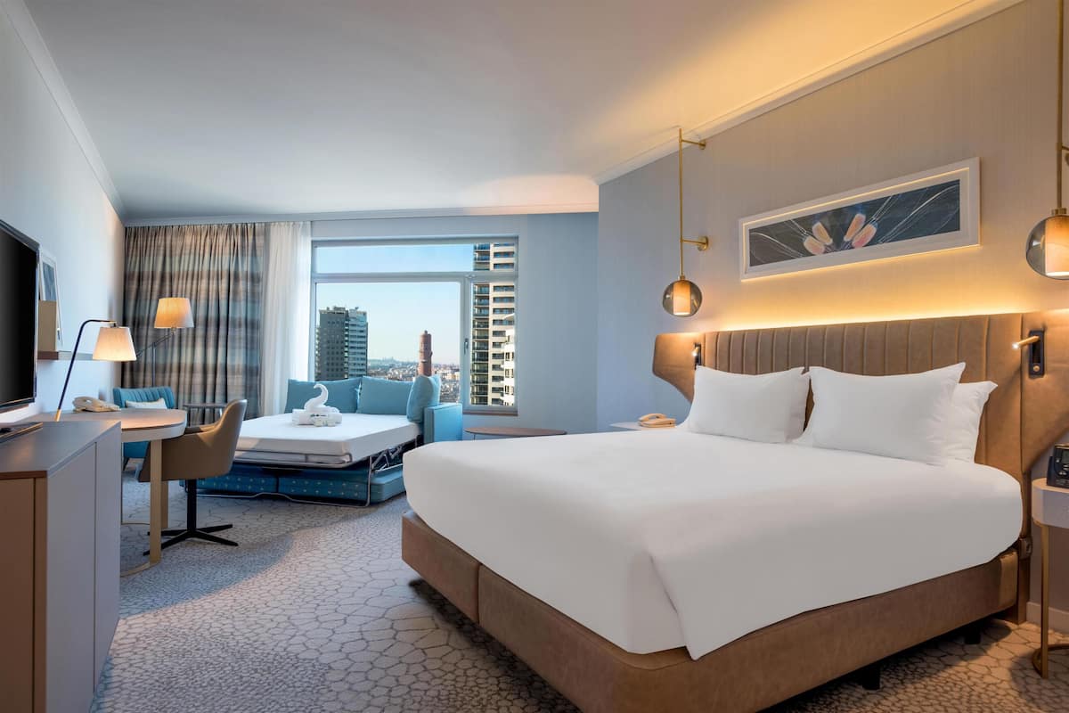 A modern hotel room with a large bed, a desk, a chair, a sofa, and a view of the cityscape.
