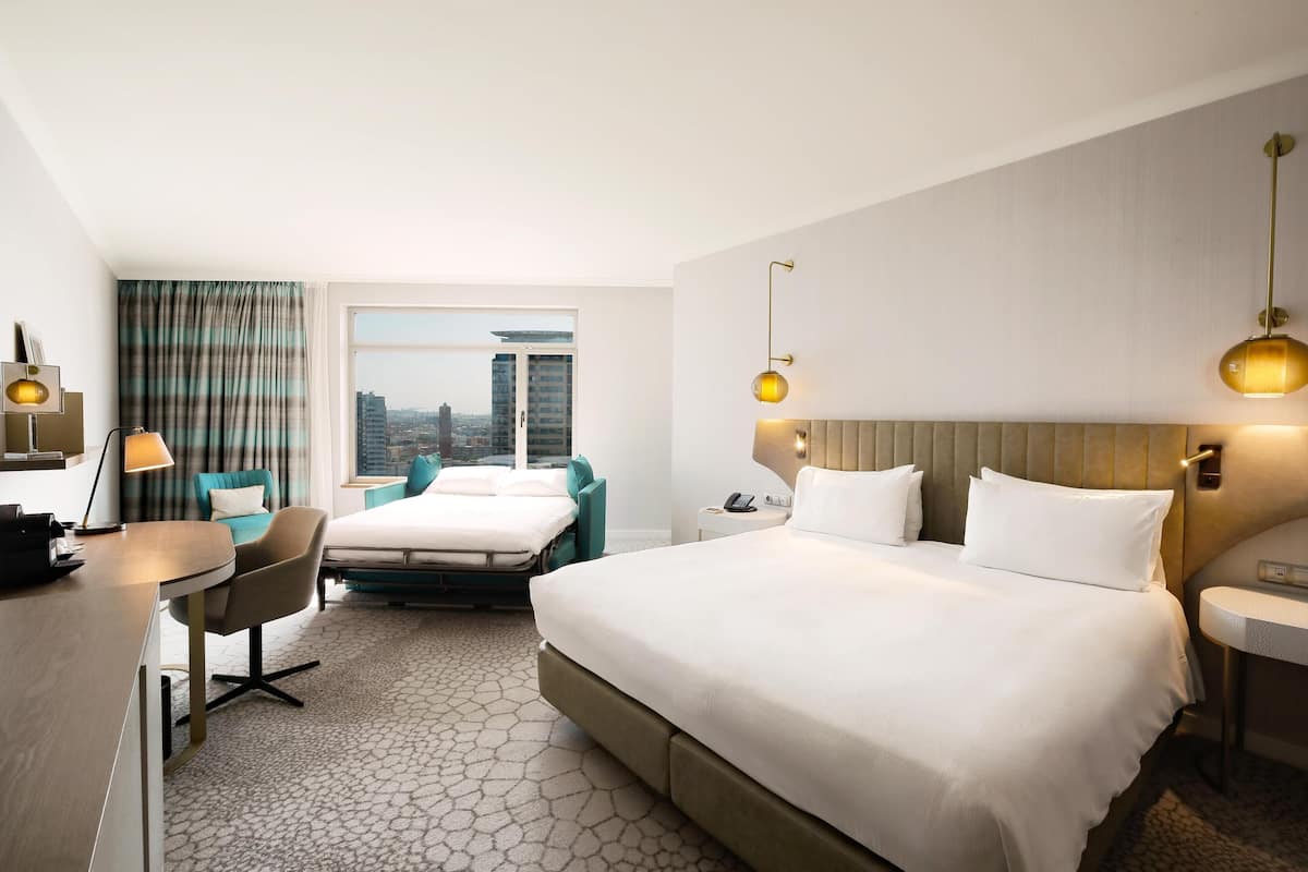 A hotel room with a large bed, a desk, a chair, and a view of the city through a large window.