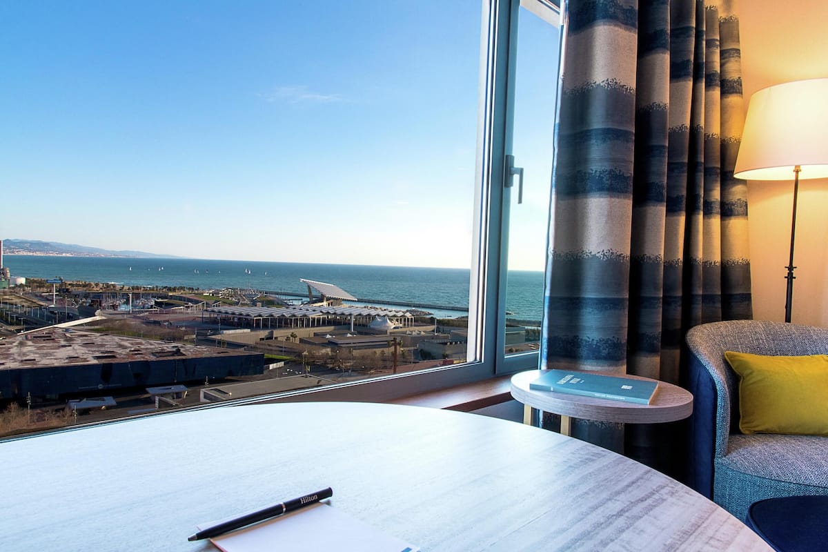 A hotel room with a large window offering a view of the sea and a distant cityscape.