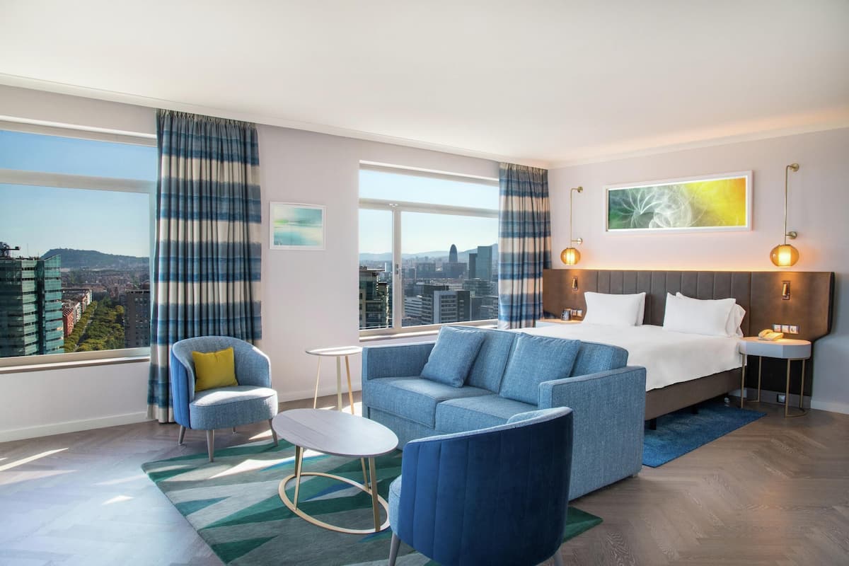 A modern hotel room with a large bed, a blue sofa, a small table, and a chair. There are large windows with curtains, a city view, and a painting on the wall.