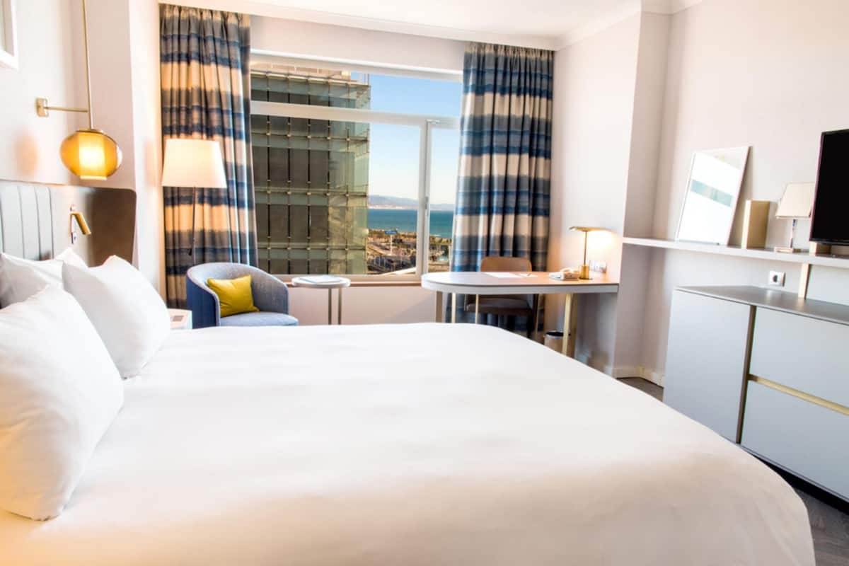A hotel room with a large bed, a small table, a chair, a TV, and a view of the city through the window.